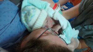 Le bébé est tenu par une infirmière, devant la maman qui l'entoure dún bras. Bébé a un bonnet et est couvert dans une couverture.