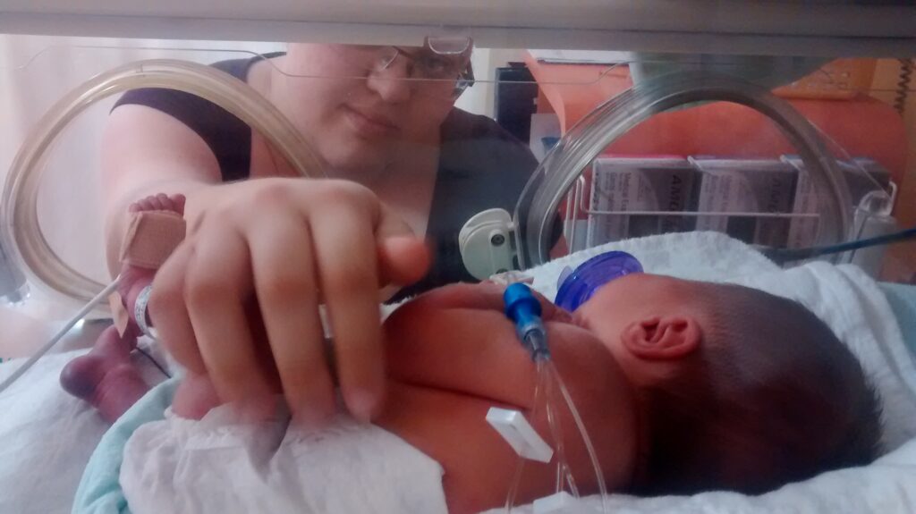 Bébé prématuré de dos, tourné vers sa maman qui a mis sa main dans l'incubateur pour le toucher.